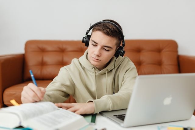German student wearing headphones and taking notes beside a laptop.