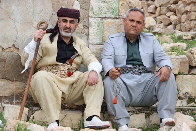 1. Two Kurdish men wearing traditional clothing and sitting beside a stone wall.